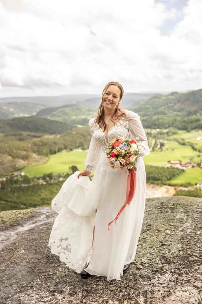 Astrid i brudekjole med blomsterbuket på fjellknaus, naturskjønt landskap i bakgrunnen.